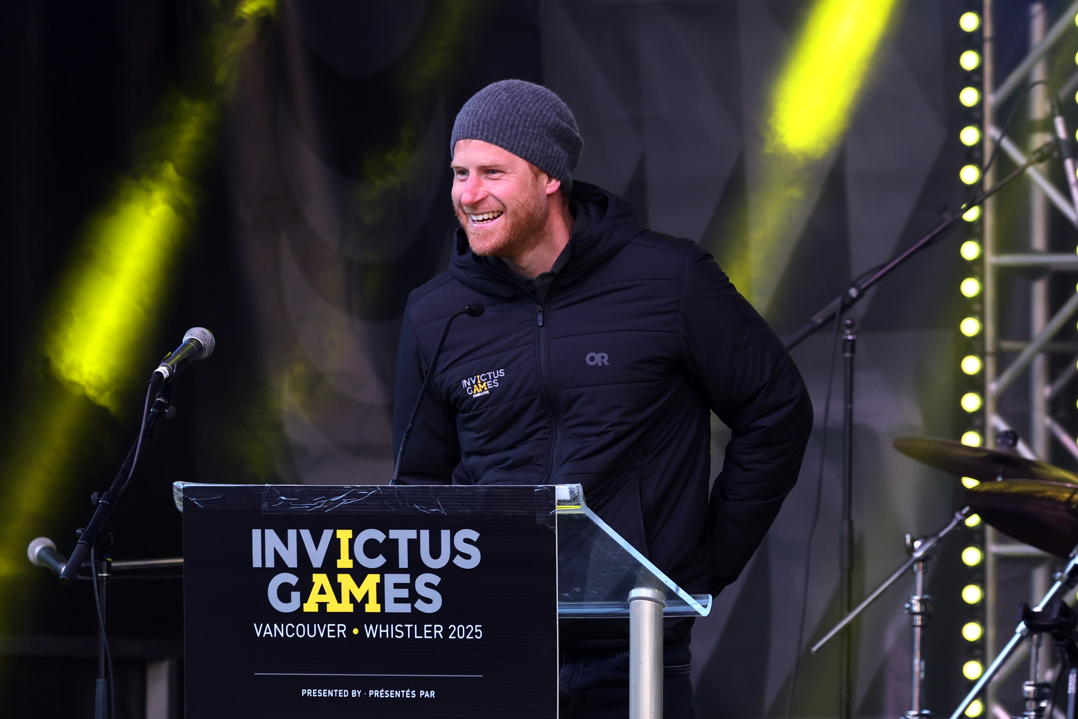 Prince harry standing on stage wearing a black coat and gray hat standing at a podium that says Invictus Games Vancouver Whistler 2025