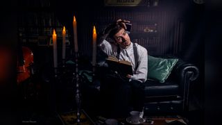 Person in a vintage room holding a book on a leather sofa, with candles and a violin nearby