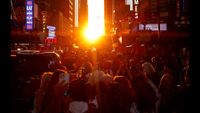 The sun peeks between tall buildings as a crowd forms underneath. 