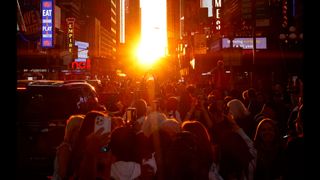 The sun peeks between tall buildings as a crowd forms underneath. 