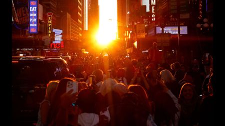 The sun peeks between tall buildings as a crowd forms underneath. 
