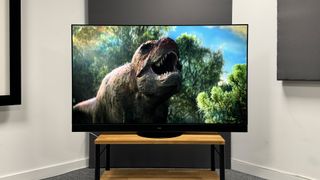 The Panasonic MZ2000 OLED TV, photographed against a white and grey background. On the screen is an image of a roaring dinosaur.
