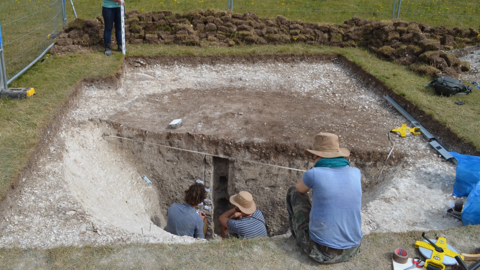 In this image we see a large square piece of grass that has been dug up to reveal a 10,000-year-old pit. Inside the pit are 2 archaeologists taking samples. There are 2 more archaeologists outside the pit, on opposite sides, taking measurements. Several tools are strewn about the site.
