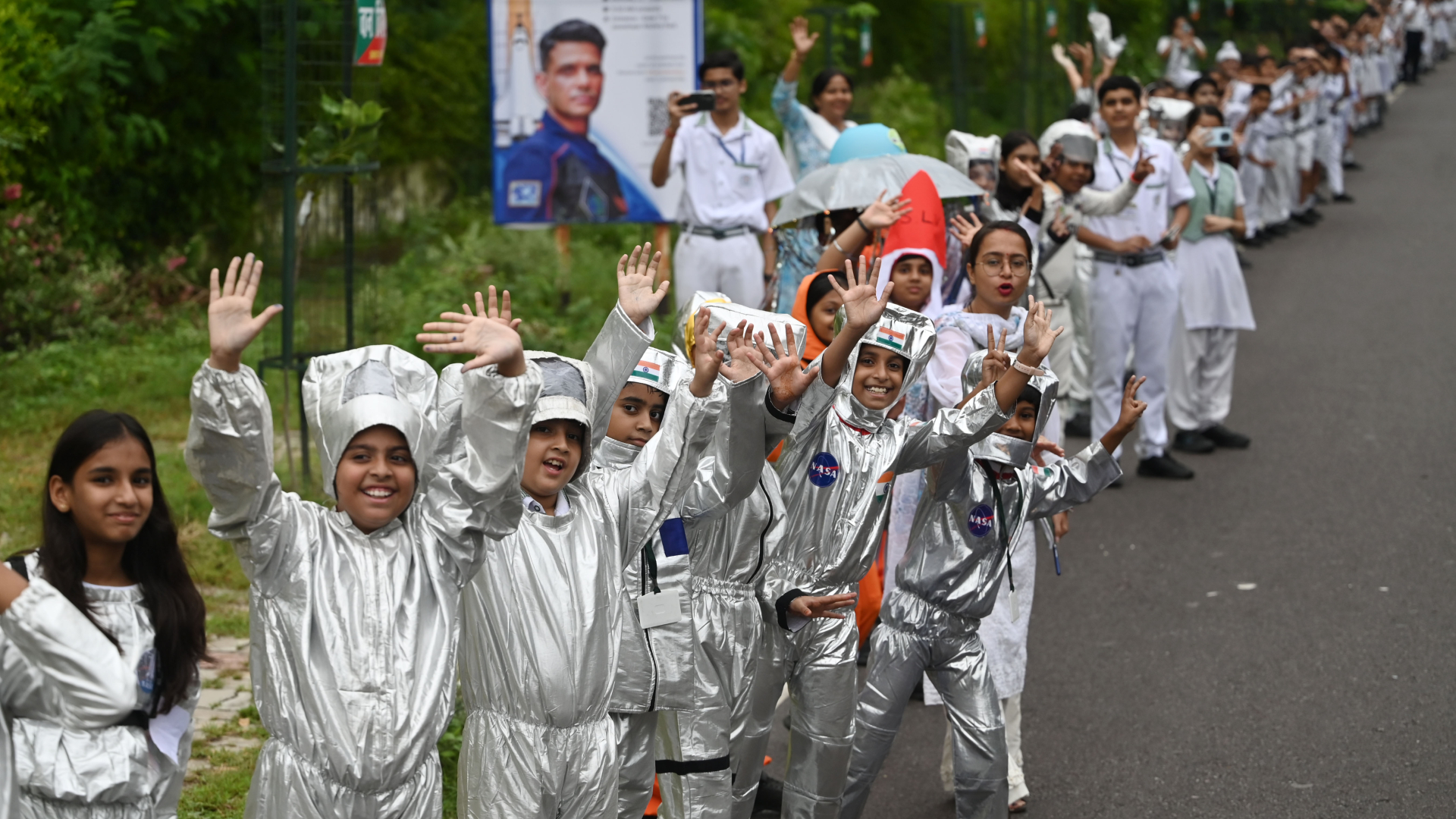 Small children wearing shiny silver outfits line a side of the street and wave to the camera