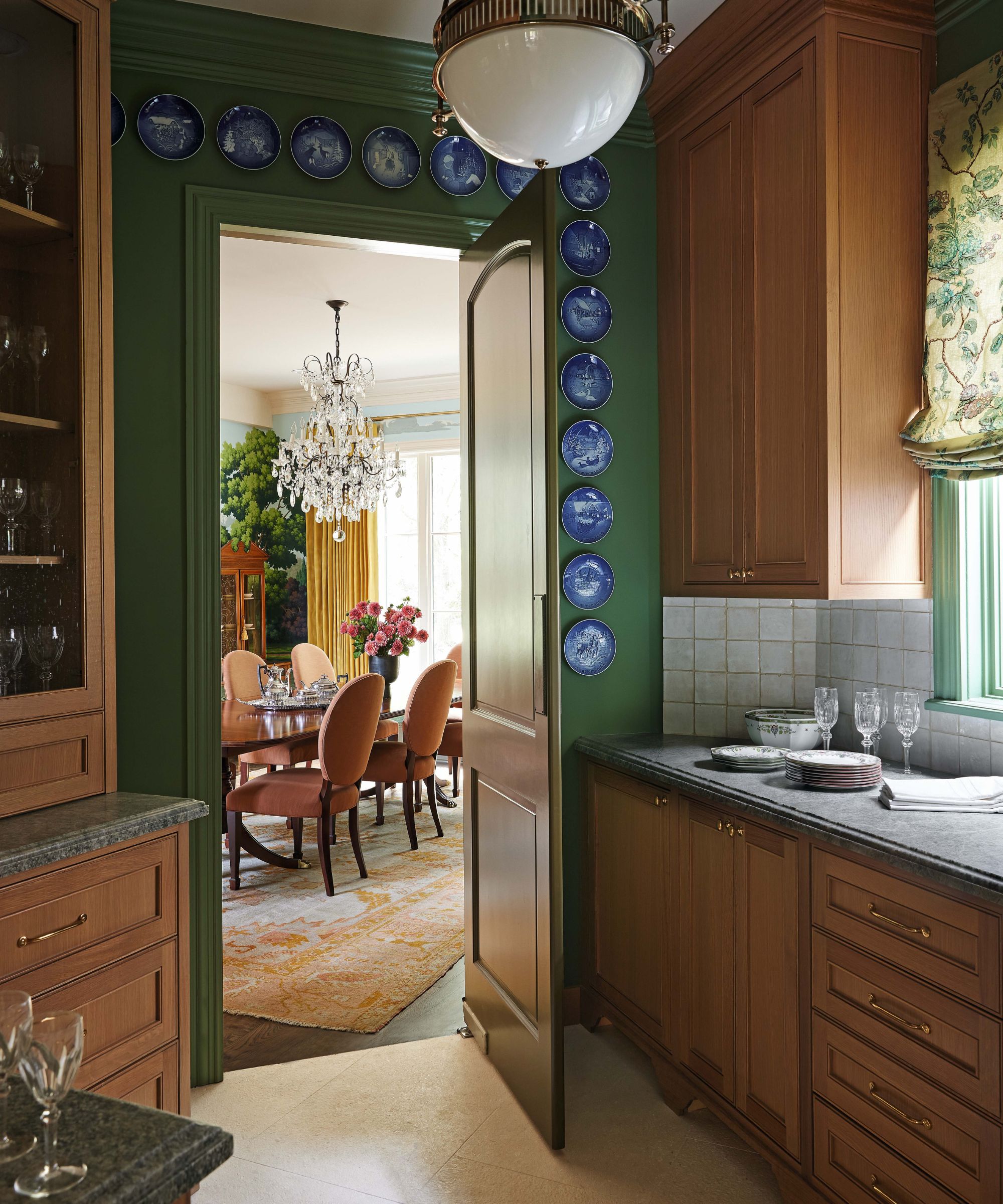 A view from a dark green butler's pantry with wood cabinets and decorative blue plates on the wall, looking through an open door into a formal dining room with a crystal chandelier