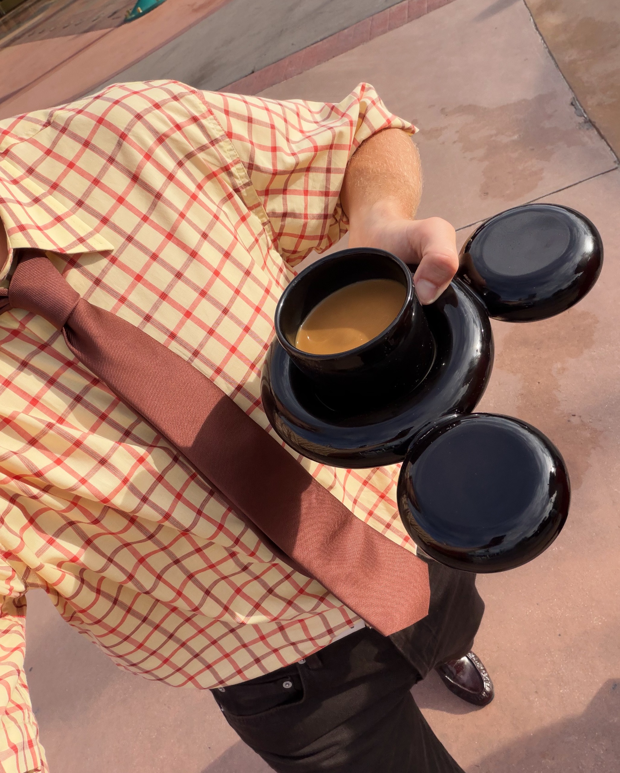 A person in a checkered beige and red shirt, brown tie, and black trousers holds a mouse-shaped, three-part glazed black ceramic mug filled with tea.