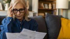 An older woman in glasses reviews her bills while sitting on the couch.