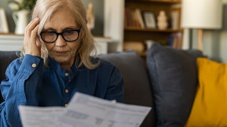 An older woman in glasses reviews her bills while sitting on the couch.