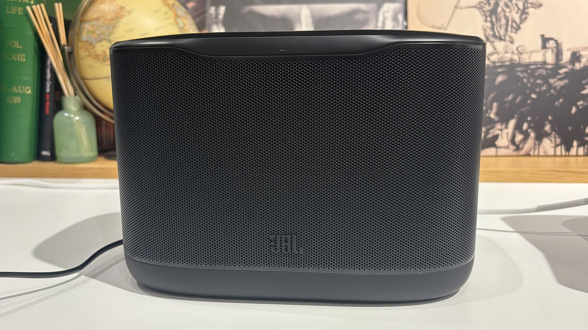 JBL Cove X1 multi-room wireless speaker on white table in front of busy bookshelf
