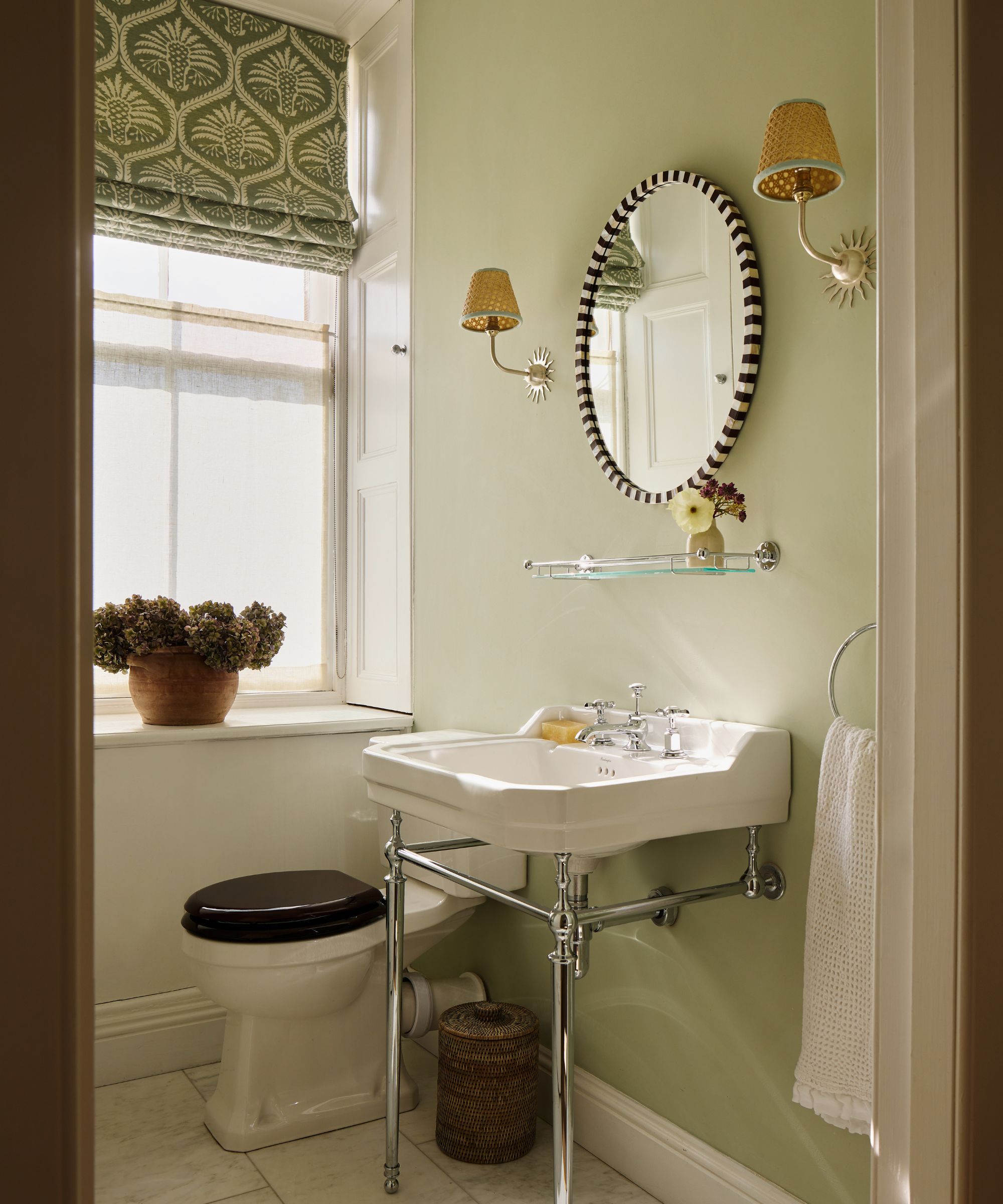 Charming sage green bathroom with a vintage-style chrome-legged sink, an oval striped mirror, and a green patterned window shade