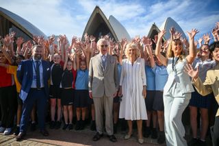 Queen Camilla and King charles standing in front of Sydney Opera House with a group of people waving their arms in the air