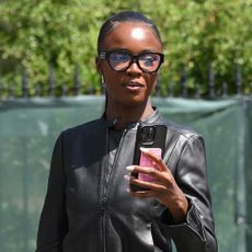 Leomie Anderson wearing a black leather jacket, black reading glasses, and holding an iPhone in London