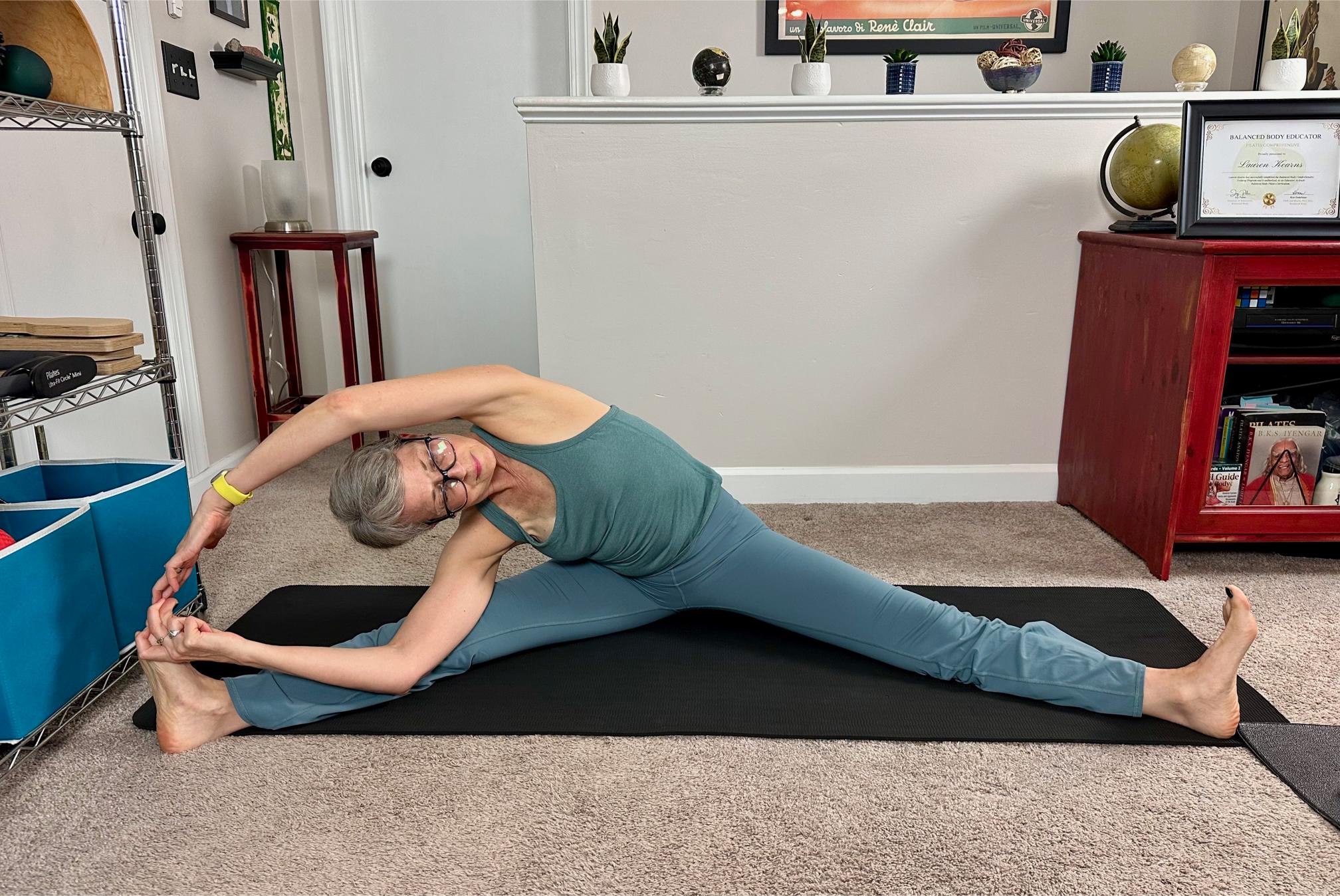 woman seated on the floor on a black yoga mat, legs wide stretching to the side
