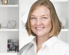 a headshot of certified professional organizer Kimberly Corey standing in front of a white shelf unit with well-space objects on it: a lady with a blonde bob in a white long sleeved collared shirt