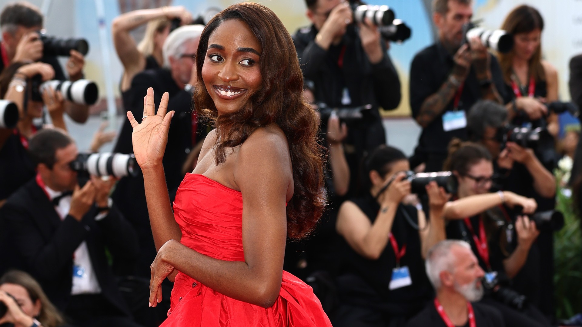 a black woman with long brown hair wearing a strapless red gown on a red carpet in front of a crowd of paparazzi