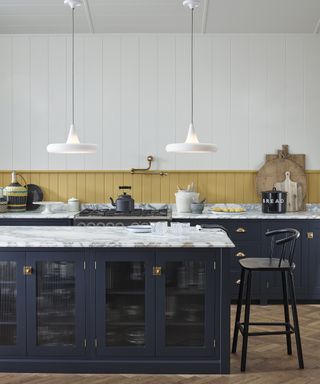 A kitchen with blue island, marble island worktop and shiplap walls painted in white and yellow with two white pendant ceiling lights over island
