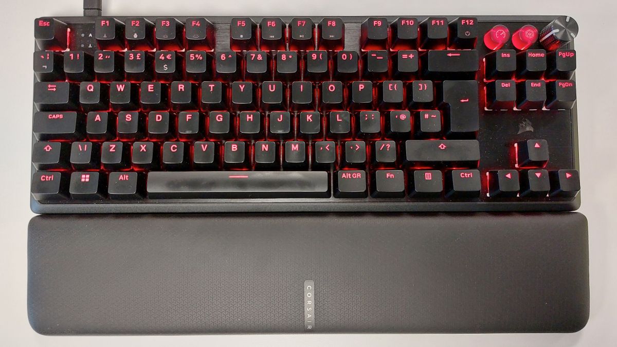 The Corsair K70 Pro TKL gaming keyboard seen from above, with the wrist rest attached, on a well-lit desk. Game mode has been activated, bathing every key in red light.