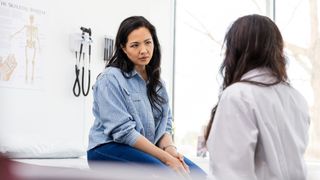 A female patient is sat on a bed in a clinic while a doctor (who is facing towards the patient but away from the camera) talks to her.