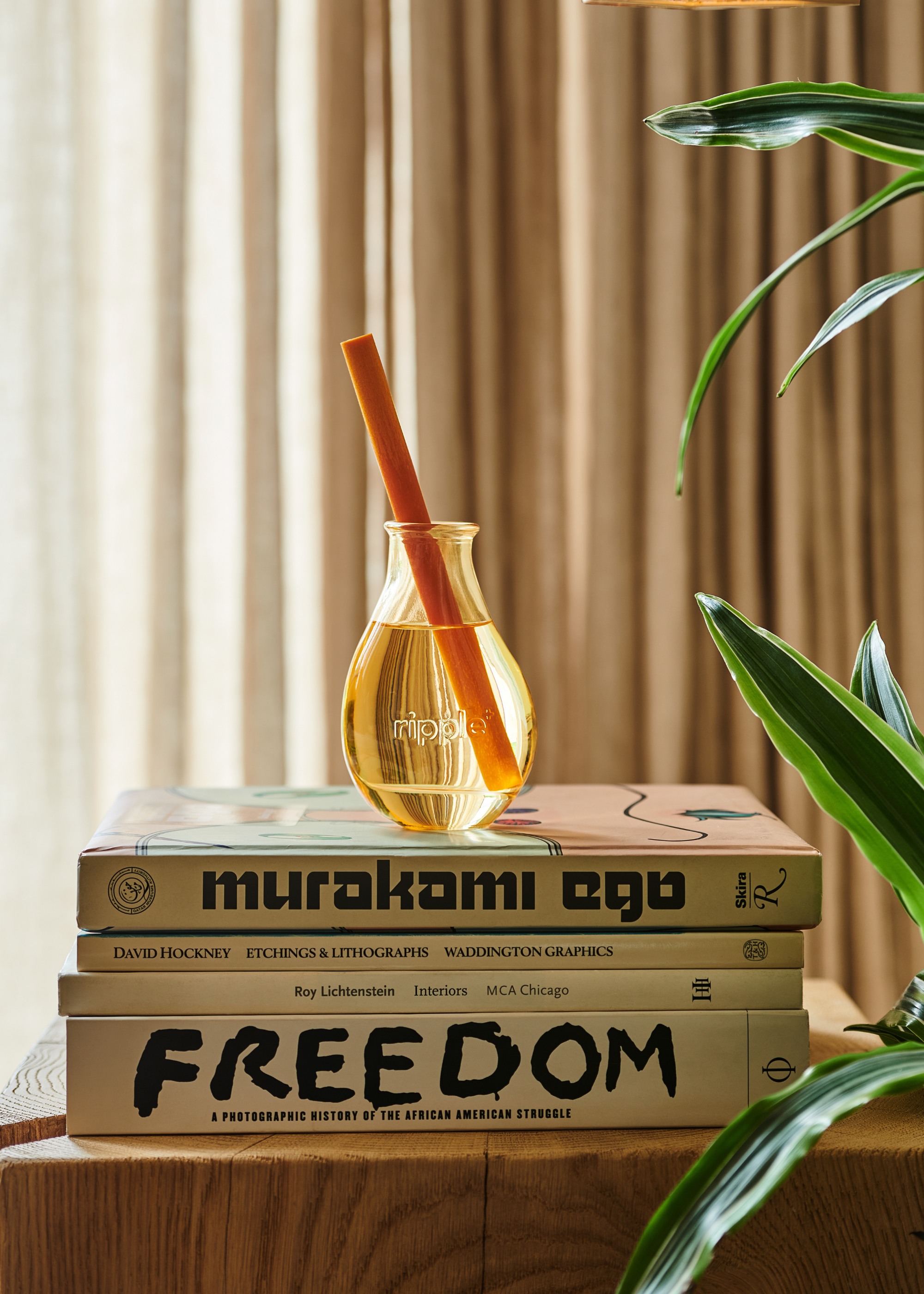 A yellow glass reed diffuser on a stack of books, on a wood plinth, by a potted plant