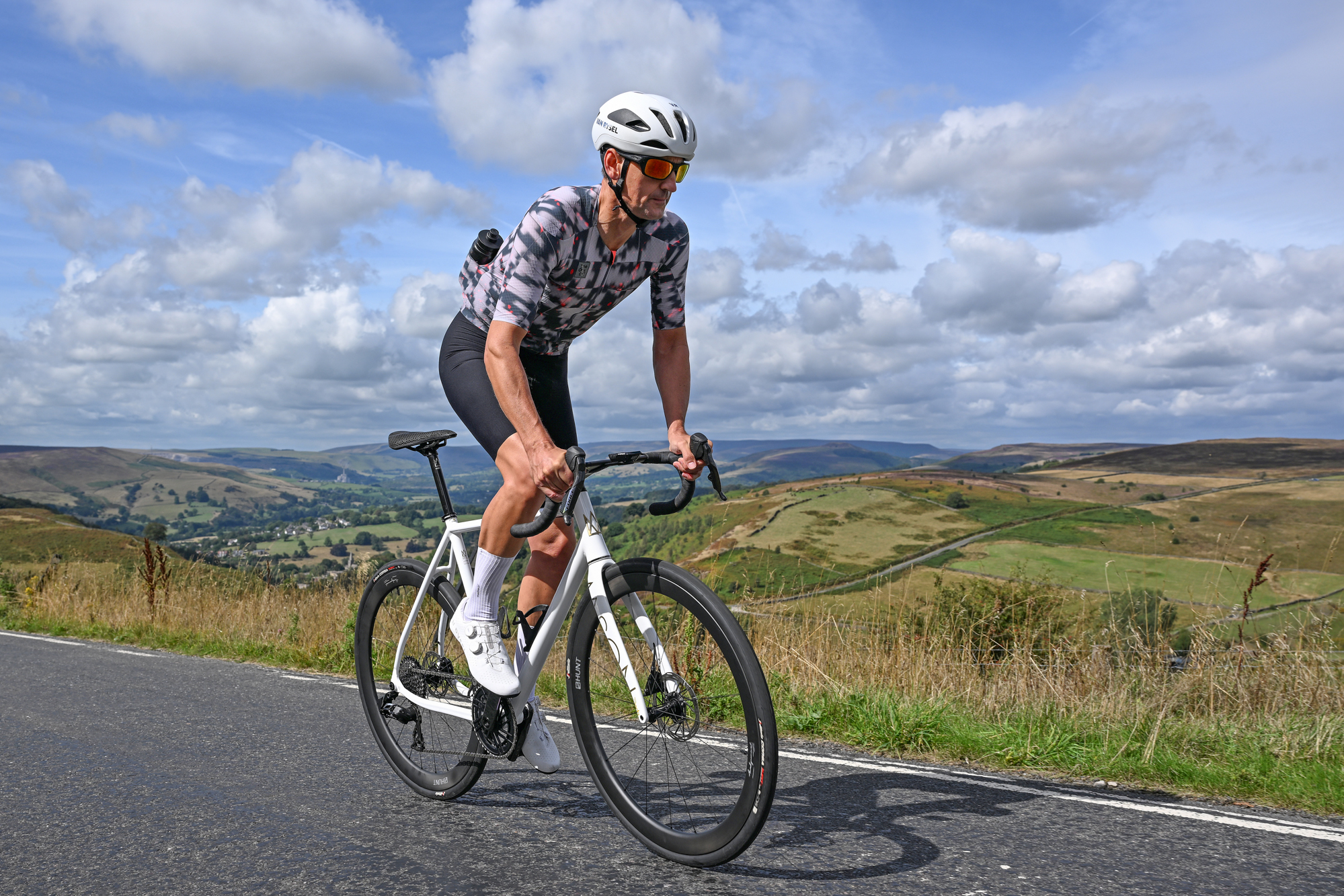 a white road bike being ridden out of the saddle uphill by a man wearing white shoes, white helmet, black shorts and a patterned grey jersey with countryside hills and blue sky in the background