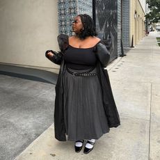 Chichi wears long gray pleated skirt, black puff sleeve top, faux leather trench, black slingbacks with a white sock. 