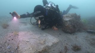 Here we see a scuba diver underwater using a flashlight to illuminate a gravestone found on the seabed.