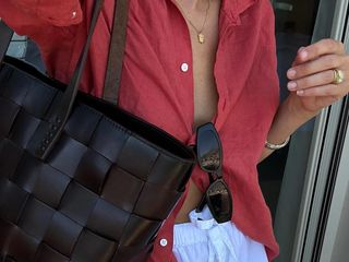 British fashion influencer Lucy Williams poses in a summer outfit with a simple gold pendant necklace, red linen button-down shirt, black sunglasses, black woven Dragon Diffusion bag, and white cotton drawstring shorts