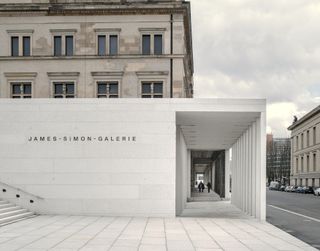 The James-Simon-Galerie at street level. The name appears on a white block wall and the side-walk is covered.