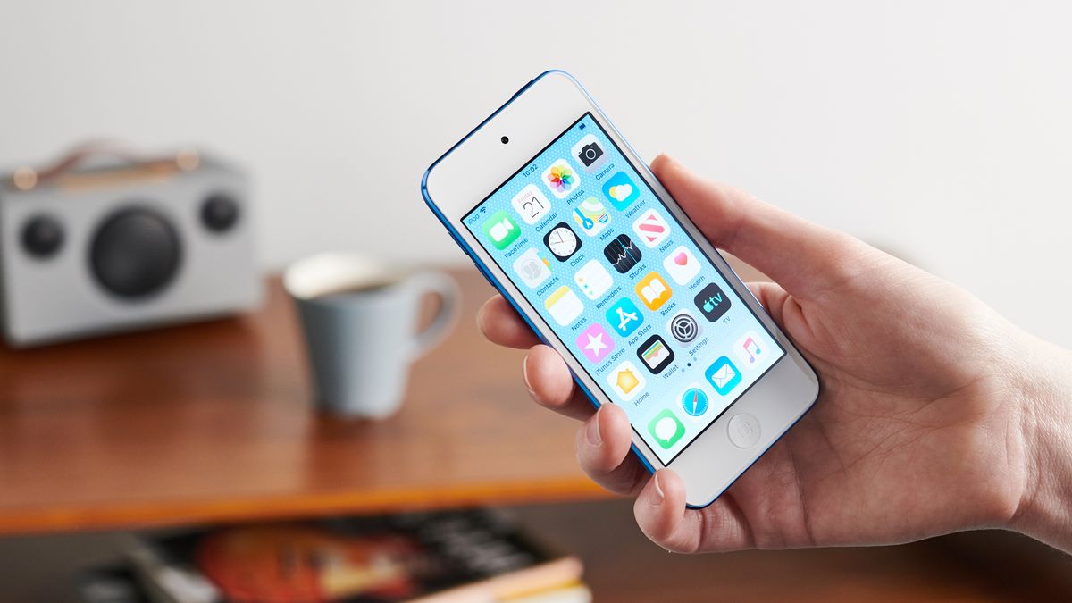 A person holding a blue Apple iPod touch (7th generation) looking at the apps on the home screen with a mug and a radio in the background on a wooden surface