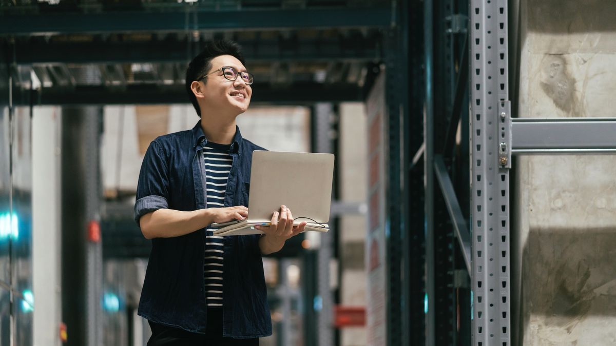 A man in a warehouse, holding a laptop, while looking at stacked shelves