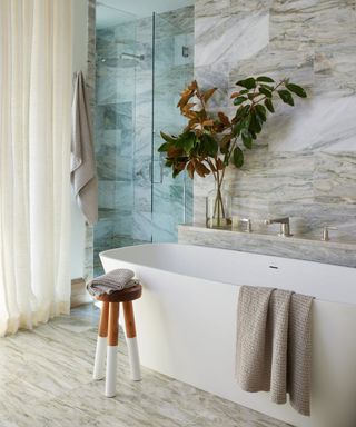 modern, minimalist marble bathroom with white bath, wooden stool, vase of flowers, cream curtains and gray towels