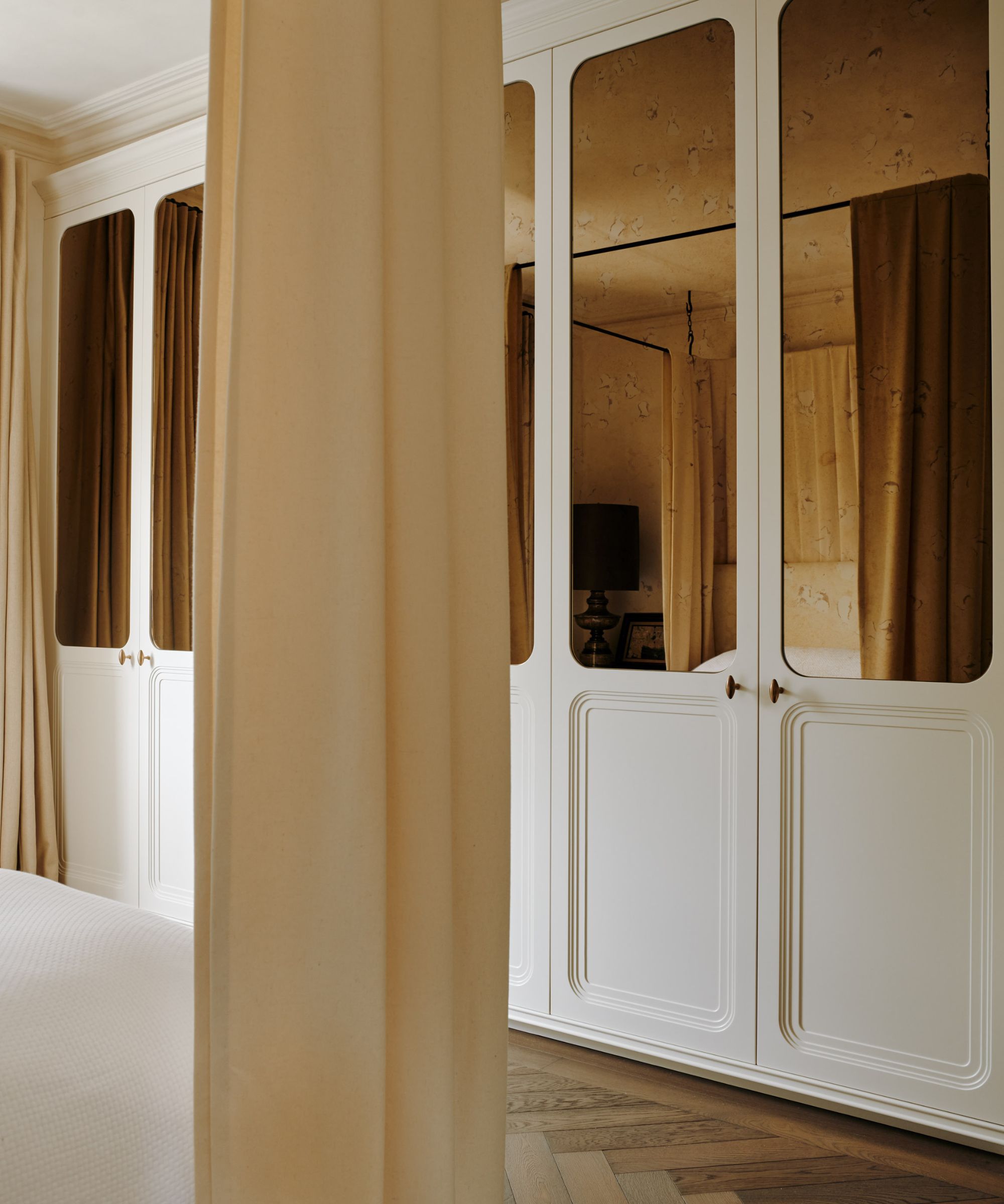 Bespoke built-in white wardrobes with mirrors in bedroom, dark wood floor and neutral curtains