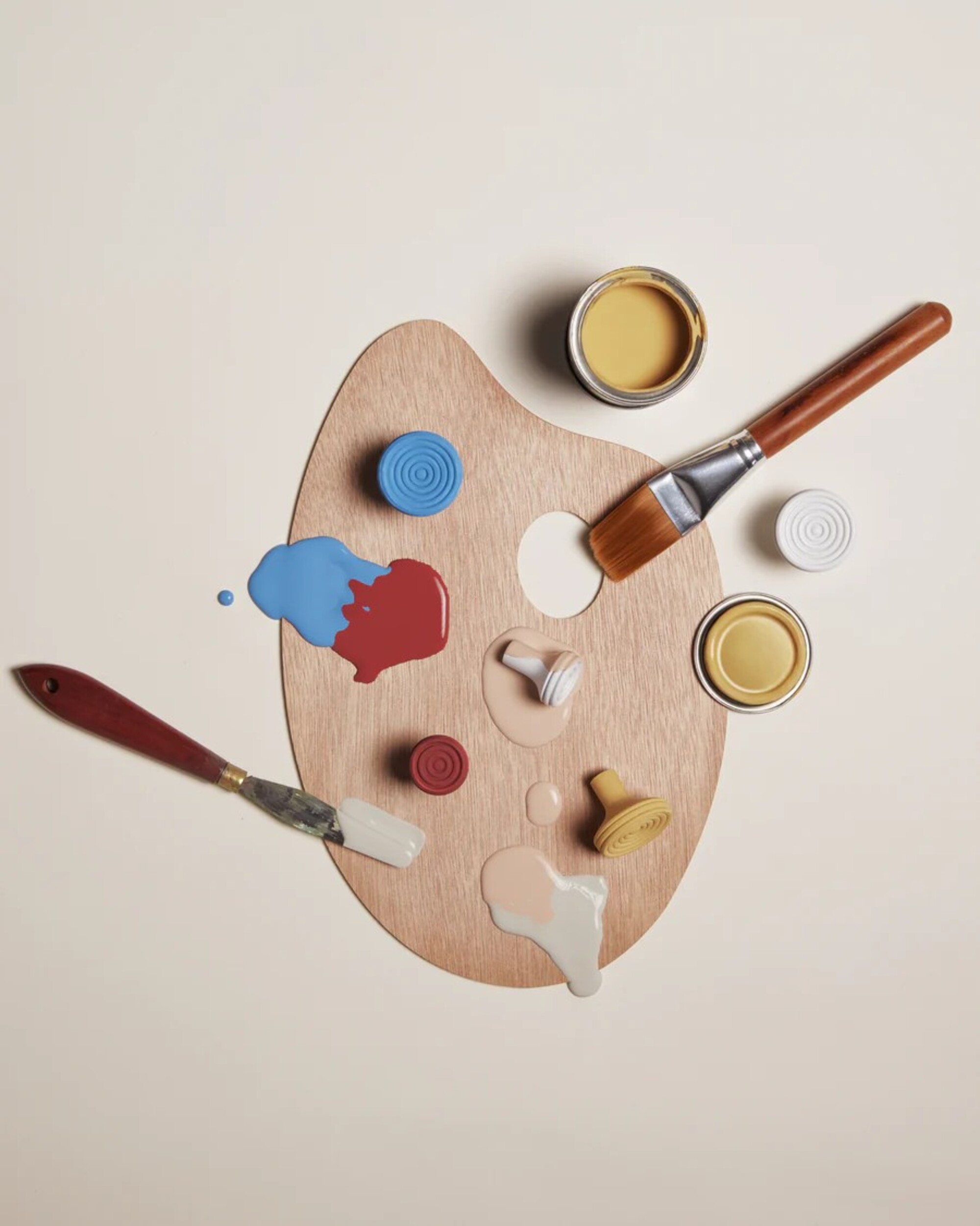 A wooden paint palette with spilled paint on it, two paint brushes, an open can of yellow paint, and three different colored doorknobs.