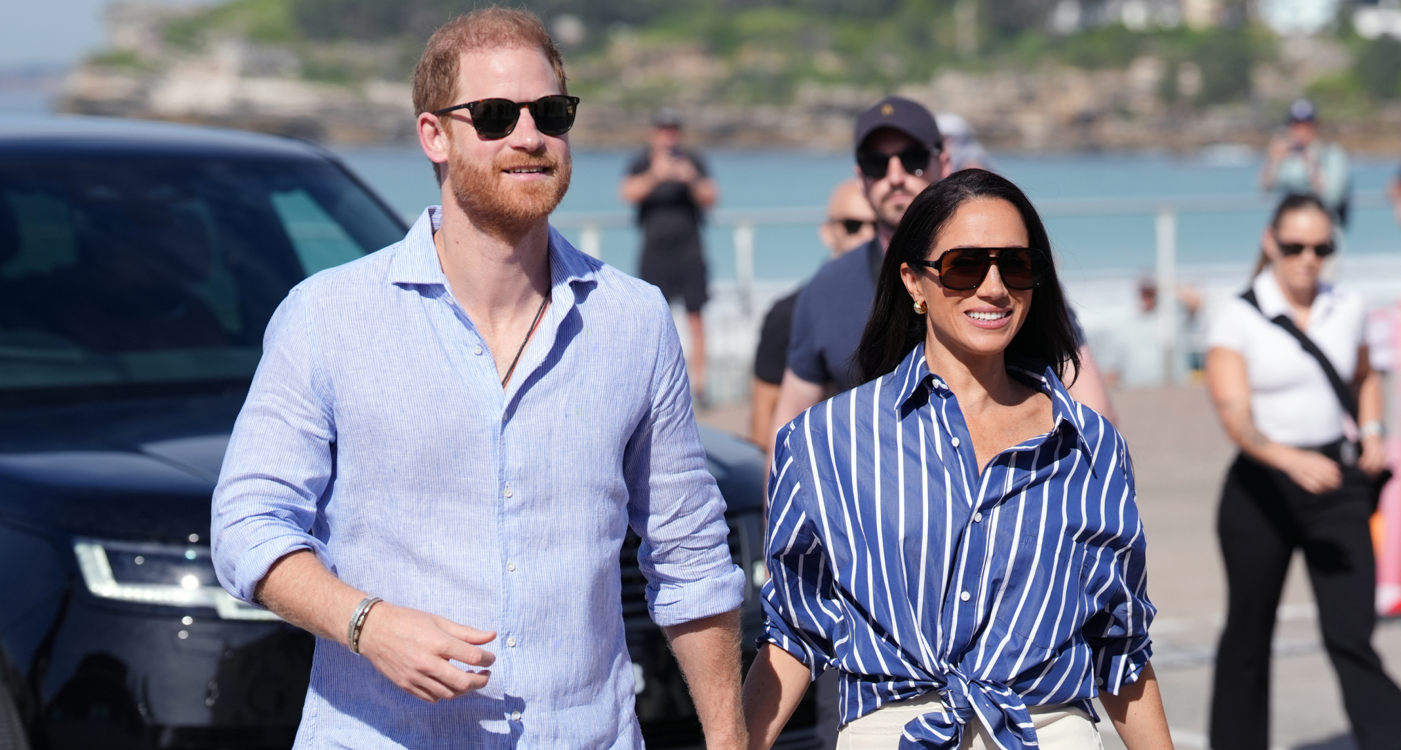 Prince Harry and Meghan Markle wearing sunglasses and blue shirts walking outside