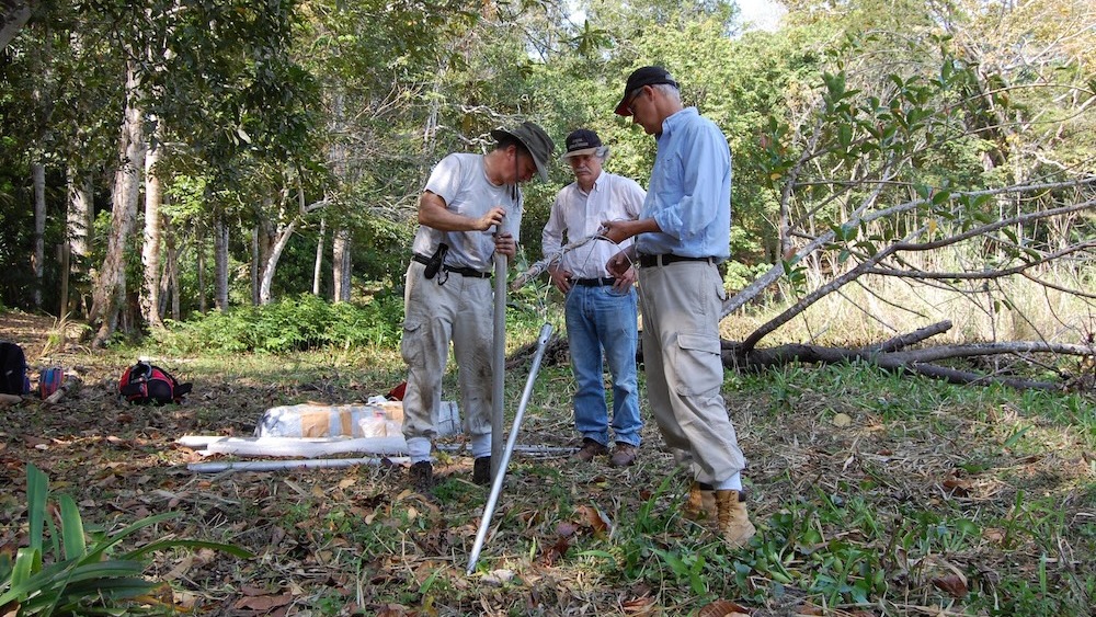 Three men stand in a forest during an archaeological dig