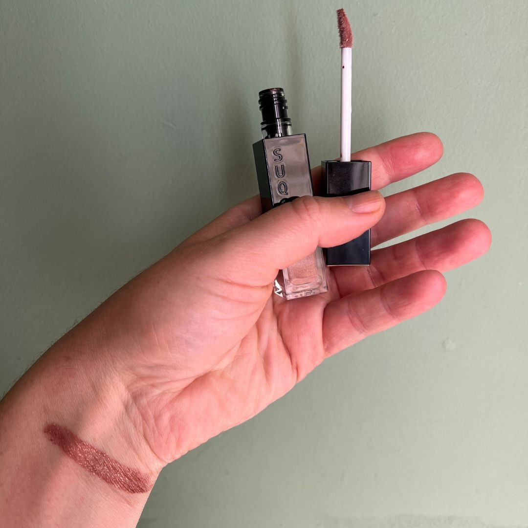 a hand on a green background holding x cream eyeshadow with a swatch of the eyeshadow on the wrist 