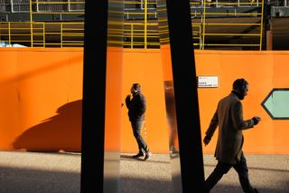 Photograph taken with Fujifilm X100V, pedestrians walking against orange hoarding background