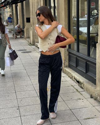 French style influencer Anne Laure Mais poses on a Paris sidewalk in a brown vest, Adidas sweatpants and mesh ballet flats