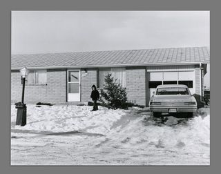 Black and white photograph showing the front of a house with a car on the drive, the drive and garden is covered in snow, and a small child is standing in the snow