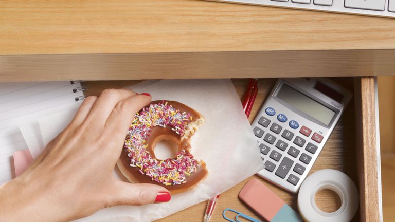 Woman putting bitten doughnut in desk drawer with stationery