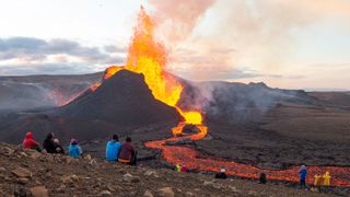 A group of people watch from afar as a volcano explodes with lava