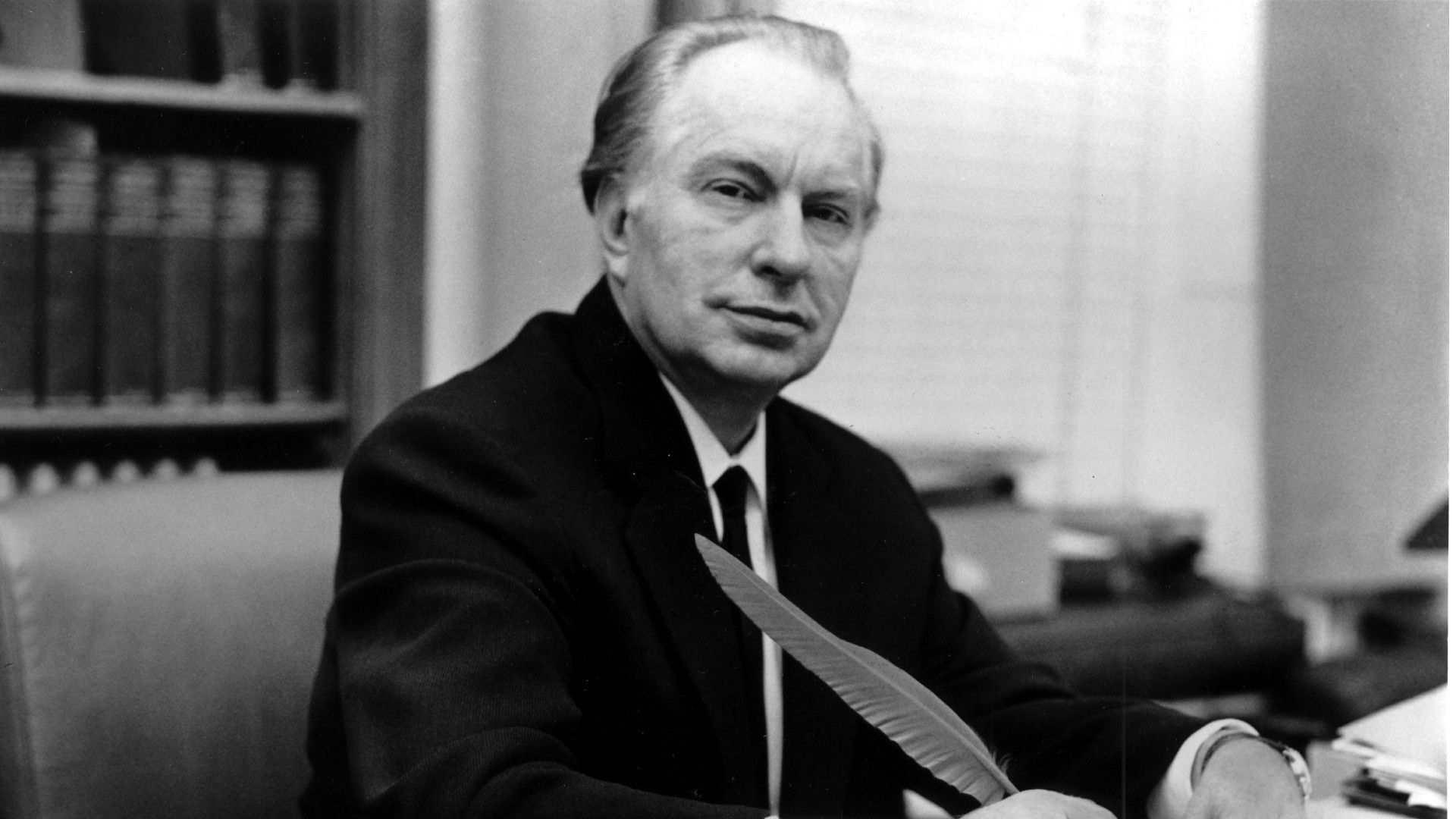 a man in a suit sits at a desk, holding a feather quill pen