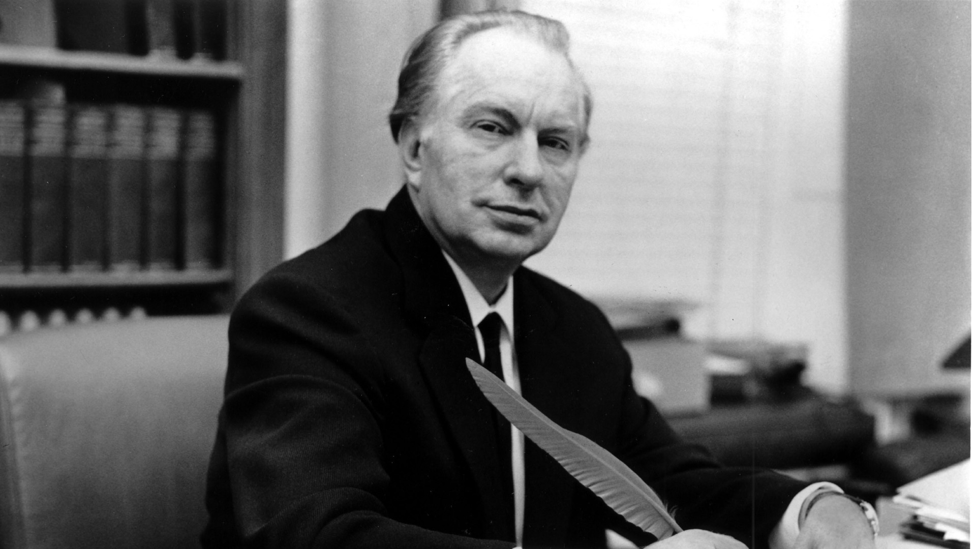 a man in a suit sits at a desk, holding a feather quill pen