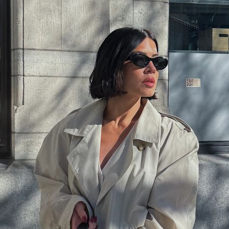 A woman with a fine hair bob hairstyle, wearing sunglasses and a trench coat