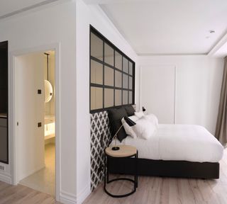 A wider view of a room at The Serras featuring wood flooring and white walls with a section behind the bed that has a black and white diamond pattern. The bed has black and white pillows and linen and there is a black grid style window above the bed. There is also a round wooden and black side table with a lamp and a partial view of another room