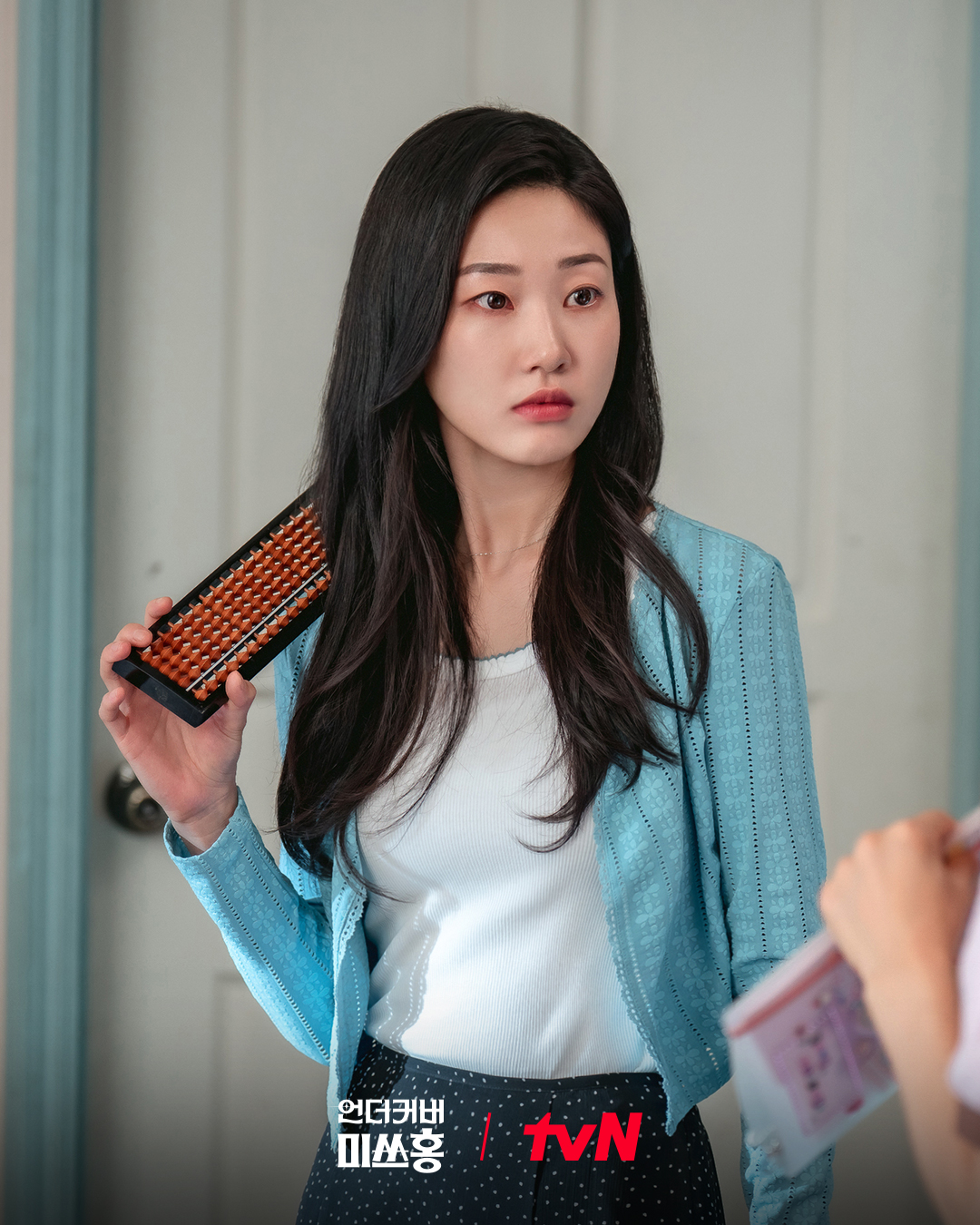A Korean woman wearing a white shirt and blue cardigan (Ha Yoon-kyung as Go Bok-hee) rests an abacus on her shoulder while standing in front of a door, in the Netflix K-drama 'Undercover Miss Hong.'