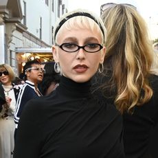 Emma Chamberlain wearing a black longsleeve top, black skirt, black sheer tights, and black pointed toe shoes