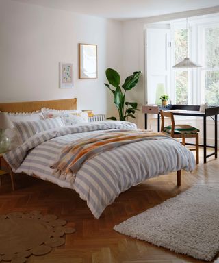 A bedroom office with blue and white striped duvet, scalloped pillowcases, small jute rug, and wooden desk facing window with large indoor plant