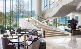 Interior view of Kerry Hotel, Hong Kong, China featuring light coloured flooring, floor-to-ceiling windows, a white staircase with glass balustrades, grey chairs, round tables and curved seating