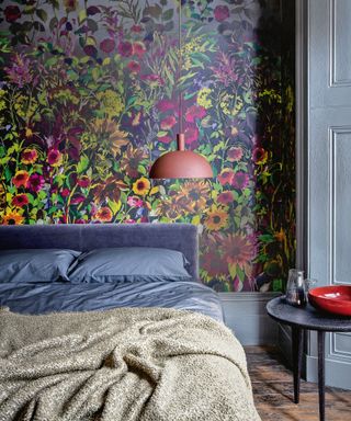 Dark gray painted bedroom with multi-color floral wallpaper behind bed, blue-gray textured headboard with dark gray bedding, rust red hanging pendant, dark wooden flooring with black side table